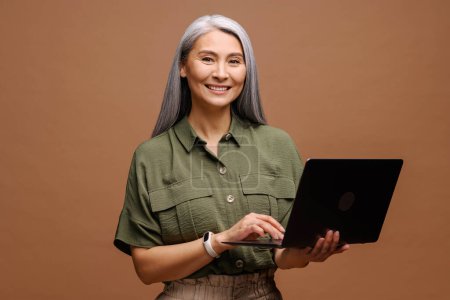 Foto de Gray-haired 40s 50s Empresaria asiática usando computadora portátil aislada, estudiante de mediana edad escribiendo, mujer alegre respondiendo a correos electrónicos, concepto de publicidad - Imagen libre de derechos