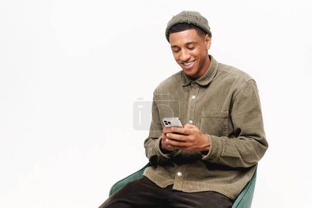 Photo for Joyful and carefree African-American 20s man using smartphone sitting in armchair isolated on white, guy in hat spends leisure in social networks, chatting online, shopping, using new mobile app - Royalty Free Image