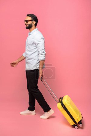 Photo for Vertical full length view at handsome middle-aged Indian bearded man wearing sunglasses walking with luggage yellow suitcase on pink background, goes on vacation - Royalty Free Image