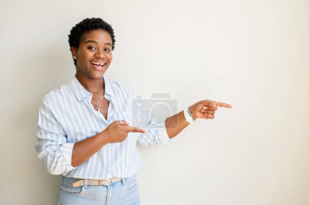 Téléchargez les photos : Femme africaine élégante avec les cheveux courts et l'écart entre les dents souriant et pointant les doigts de côté à l'espace de copie, montrant la zone pour la publicité. Studio intérieur shoot isolé sur fond blanc - en image libre de droit