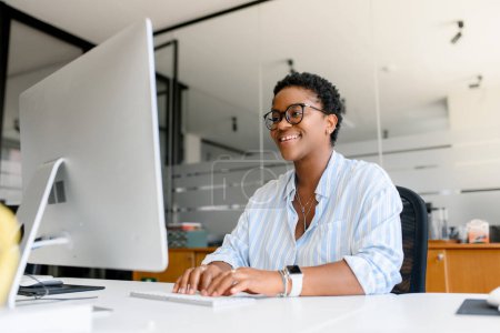 Téléchargez les photos : Femme afro-américaine réussie assise au bureau avec ordinateur portable, souriante et dactylographiant, appréciant le travail de bureau, employée, femme d'affaires se sentant au bon endroit - en image libre de droit
