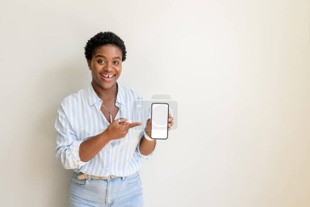 Foto de Mujer afroamericana feliz señalar con el dedo a la pantalla vacía del teléfono inteligente, la publicidad de la nueva aplicación móvil empleada de oficina femenina multiracial mirando a la cámara, espacio de copia, aislado en blanco, plantilla - Imagen libre de derechos