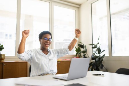 Foto de Emocionada mujer afroamericana freelancer levantando las manos en éxtasis, mujer de negocios consiguió la promoción, oferta de trabajo, celebrando el éxito, mirando a la pantalla del ordenador portátil, gritando sí, triunfo y concepto de victoria - Imagen libre de derechos
