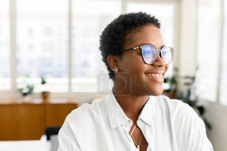 Foto de Retrato de empleada afroamericana confiada con el pelo corto parado en el espacio moderno de la oficina, mujer de negocios negra inspirada atractiva alegre que mira a un lado con sonrisa amistosa ligera - Imagen libre de derechos