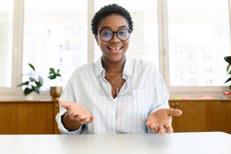 Foto de Atractiva y enérgica colega afroamericana, tutora, entrenadora hablando en línea durante una videollamada, sentada en el escritorio, mirando a la cámara y hablando con gestos. Reunión virtual, conferencia - Imagen libre de derechos