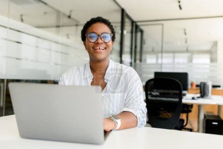 Téléchargez les photos : Employée de bureau afro-américaine moderne avec une coupe de cheveux courte portant une chemise décontractée intelligente et des lunettes, femme d'affaires heureuse assise au bureau avec un ordinateur portable et regardant à la caméra, sourire avec des dents lacunaires - en image libre de droit