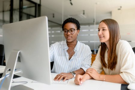 Photo for Two female colleague discussing about project sitting side by side in modern open space office, smiling and watching at computer monitor together, enjoying friendly working atmosphere - Royalty Free Image