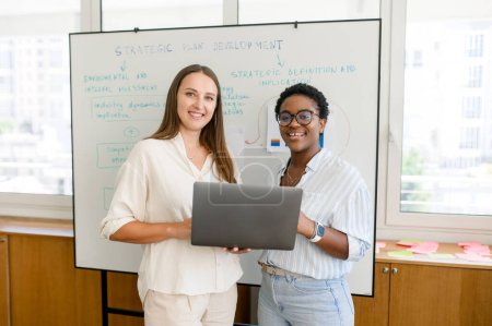 Photo for Couple of business people standing with laptop in the office. Two diverse female colleagues with whiteboard on the background in meeting room. Friendly atmosphere in team - Royalty Free Image