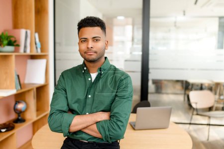 Photo for Confident Indian male employee standing with his arms folded, emanating an air of success. Freelancer working in a shared workspace, exudes a sense of determination and productivity - Royalty Free Image