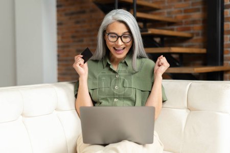 Photo for Middle-aged Asian lady looks at camera and rejoicing, made long-awaited purchase online, receive money transfer. Excited gray-haired woman holding banking card and laptop sitting on sofa at home - Royalty Free Image