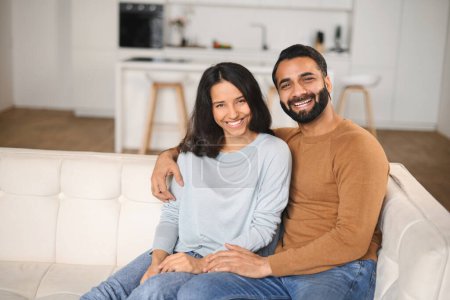 Téléchargez les photos : Gros plan d'un heureux couple interracial regardant la caméra assis sur le canapé de leur maison. Heureux propriétaires de nouveau plat souriant et embrassant devant la caméra - en image libre de droit