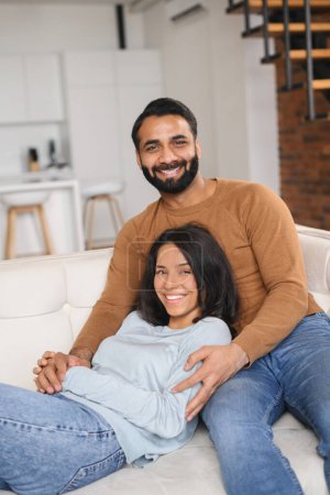 Foto de Primer plano de la vista de la pareja india de cónyuges mirando a la cámara y mostrando la forma del corazón de las manos mientras se sientan en el sofá en su nuevo piso. Foto de stock - Imagen libre de derechos