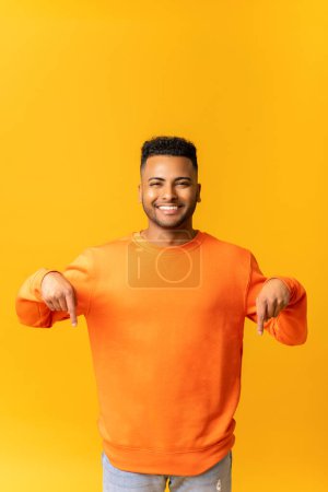 Foto de Hombre indio atractivo con expresión facial llena de alegría apuntando hacia abajo con ambos dedos, presentando espacio de copia. Estudio interior plano aislado sobre fondo naranja - Imagen libre de derechos