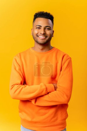 Foto de Retrato de hombre indio guapo sonriente feliz en ropa casual posando con los brazos cruzados contra el fondo naranja, chico estudiante alegre mira a la cámara con sonrisa amistosa - Imagen libre de derechos