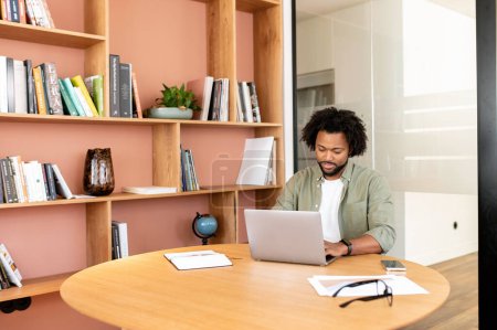 Téléchargez les photos : Étudiant afro-américain dans la bibliothèque, gars utilisant un ordinateur portable pour le travail à distance assis dans l'espace de coworking moderne, étagères en arrière-plan, - en image libre de droit