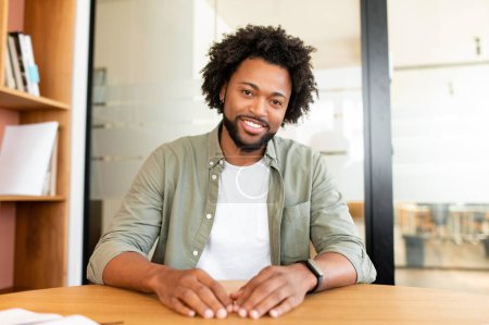 Téléchargez les photos : Portrait de beau frisé afro-américain assis au bureau et devant la caméra, vous regarde avec un sourire amical, homme d'affaires impliqué dans la vidéoconférence, participant à la vidéoconférence - en image libre de droit