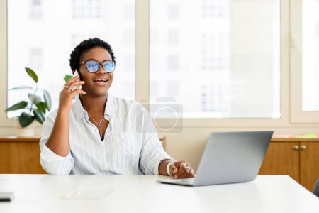 Foto de Alegre mujer de negocios afroamericana, gerente de oficina femenina en gafas que trabajan en la oficina, sentada en el escritorio con computadora portátil, sosteniendo el teléfono inteligente, hablando con el cliente o socios de negocios - Imagen libre de derechos