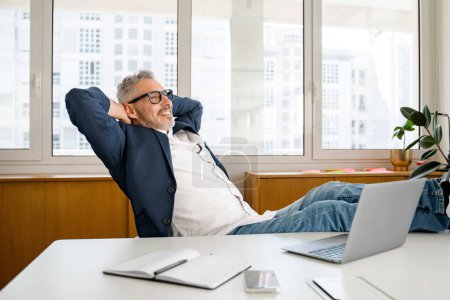 Photo for Satisfied with well done project mature gray-haired 60s businessman sits in a confident pose with his feet on the table and relaxes, leaned back in the chair, holding his hands behind his head - Royalty Free Image