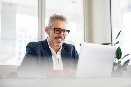 Foto de Hombre de negocios maduro sonriente usando traje formal y gafas usando el ordenador portátil para el trabajo de oficina, empleado masculino mirando a la pantalla, leyendo noticias, chequeando el correo - Imagen libre de derechos