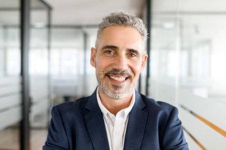 Photo for Close-up portrait of confident mature grey-haired 50s male employee standing in modern office space, cheerful experienced businessman in suit looks at the camera with light friendly smile - Royalty Free Image