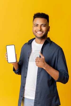Photo for Positive indian man showing thumb up and smartphone with white empty display, bragging with application. Indoor studio shot isolated on yellow background - Royalty Free Image