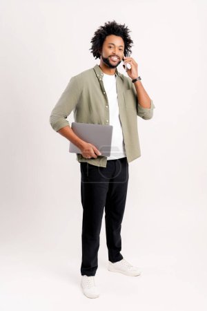 Foto de Positivo y exitoso joven freelancer en una camisa y con un portátil habla por teléfono. Concepto de estilo de vida, tecnología. Aislado sobre fondo blanco - Imagen libre de derechos