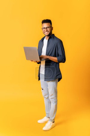 Foto de Retrato completo del empresario indio sonriente agradable usando el ordenador portátil de pie aislado en amarillo, feliz estudiante masculino determinado mirando a la cámara, la celebración de la computadora, el hombre árabe mecanografía, vertical - Imagen libre de derechos
