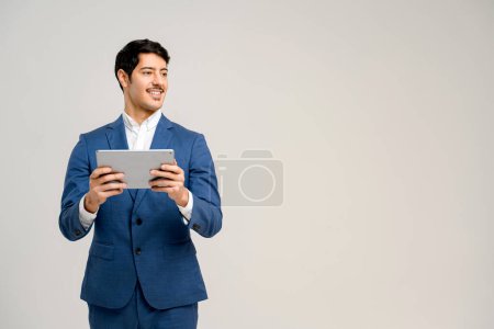 Téléchargez les photos : Le bel homme hispanique en costume formel bleu souriant tout en utilisant la tablette fournit une image relatable de l'utilisation de la technologie dans les affaires quotidiennes, le concept d'intégration de la technologie dans la vie professionnelle - en image libre de droit