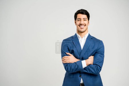 Photo for A young, confident professional man stands with arms crossed, wearing a sharp blue suit and a welcoming smile against a neutral background, representing the image of approachable modern success - Royalty Free Image
