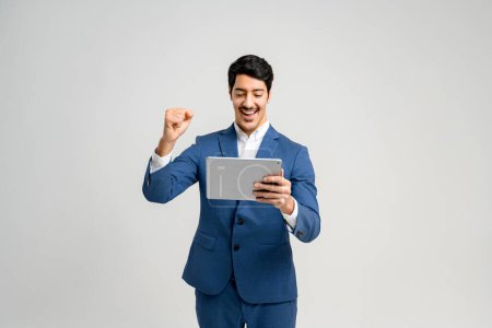 Téléchargez les photos : Homme d'affaires excité en costume bleu se réjouissant tout en regardant tablette, ce qui suggère un succès numérique ou entreprise réalisation. Concept de résultats positifs dans les entreprises numériques, négociations en ligne - en image libre de droit