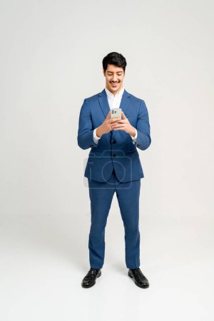 Téléchargez les photos : Portrait complet de jeune homme d'affaires confiant dans un costume bleu élégant se tient à l'aide d'un smartphone, souriant et appréciant la conversation en ligne, isolé sur fond blanc - en image libre de droit