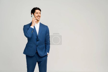 Photo for A confident businessman in a navy blue suit is engaged in a pleasant conversation on his mobile phone, portrayed against a plain background that emphasizes his professional demeanor - Royalty Free Image