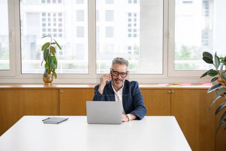 Friendly smiling mature businessman male employee has pleasant phone conversation sitting in modern office space, cheerful grey-haired boss talking on smartphone