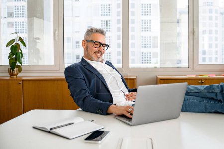 Photo for Self-assured 60s gray-haired modern confident businessman working in a comfortable and modern office in relaxed pose, putting his feet on the desk, successful mature business owner enjoying his job - Royalty Free Image