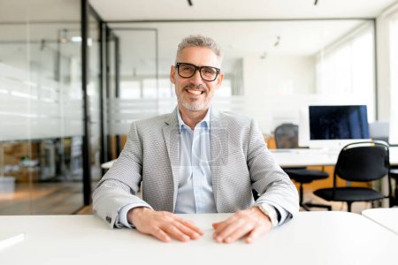 Photo for Mature businessman in a grey checkered blazer and crisp blue shirt exudes confidence as he sits at a polished office table, looking at the camera with charismatic smile - Royalty Free Image