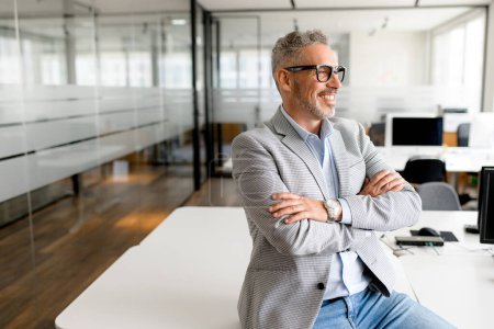 Foto de Un hombre de negocios maduro con gafas elegantes y un traje gris moderno se apoya cómodamente en su escritorio, exudando una sensación de satisfacción y logro - Imagen libre de derechos