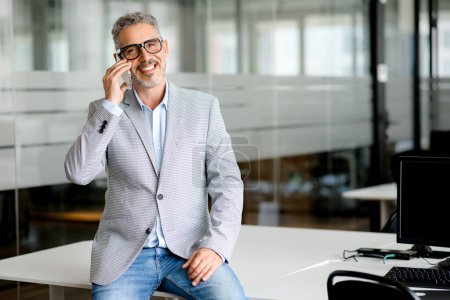 A professional middle-aged man with grey hair smiles confidently while talking on the phone in a modern office. Image captures the essence of business communication and joy of successful conversations