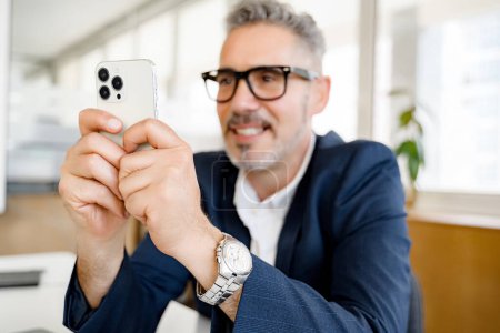 Smiling confiado hombre de negocios de pelo gris en gafas con estilo, empleado masculino utilizando un teléfono inteligente moderno, mensajes de texto, mensajería a través de conexión inalámbrica a Internet, navegación smartphone en la oficina moderna