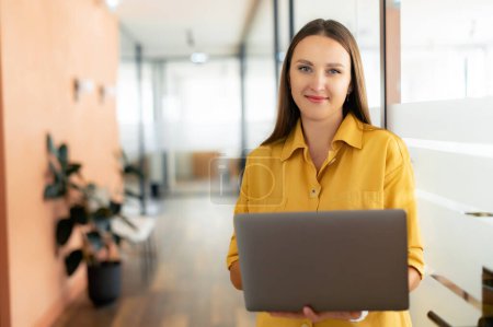 Photo for Smiling woman with laptop looks at camera and standing indoor in modern office, cheerful businesswoman in casual wear checking emails standing in hallway - Royalty Free Image