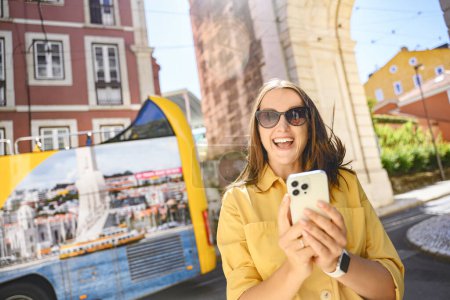 Photo for Young woman in a yellow shirt using smartphone with a tourist bus in the background. The photo showcases an interactive and playful approach to travel photography in an urban setting. - Royalty Free Image