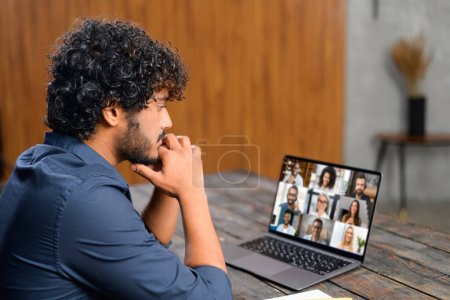Photo for Indian man sits at a wooden table, engrossed in a serious discussion during a virtual meeting with colleagues displayed on laptop screen. Focus and intensity of remote teamwork - Royalty Free Image
