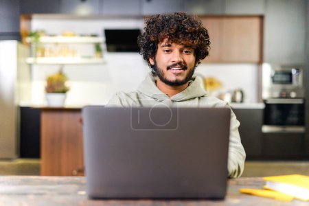 Photo for A young Indian freelancer man with curly hair is smiling while working on his laptop in a modern kitchen, exuding a vibe of casual professionalism. Positive and productive remote work concept. - Royalty Free Image