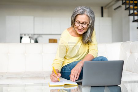 Photo for Focused retired woman is learning online, using a laptop for watching online classes, webinar, senior female takes a notes sitting in the kitchen at home - Royalty Free Image