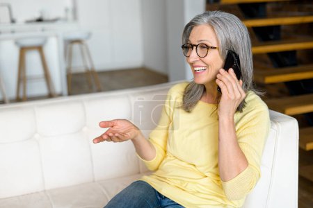 Photo for Cheerful gray-haired woman glad to talking on smartphone sitting on the sofa at home, modern senior grandmother had phone conversation, smiling and enjoying chatting - Royalty Free Image