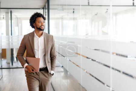 Photo for The African-American businessman is pictured in stride, holding a laptop, suggesting a moment of connectivity and the seamless integration of technology into business - Royalty Free Image