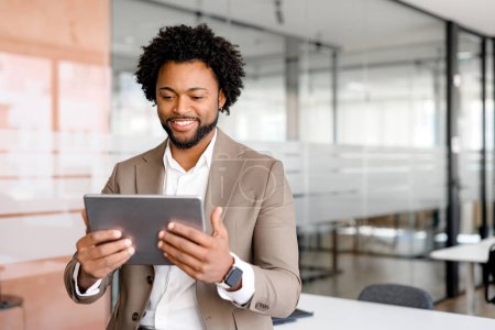 Téléchargez les photos : Un jeune homme d'affaires confiant en costume bronzé sourit tout en tenant une tablette, debout dans un environnement de bureau moderne. Concept des thèmes de l'entreprise, de la technologie et de l'entrepreneuriat. - en image libre de droit