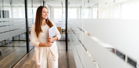 Eine junge Geschäftsfrau in einem modernen Büroambiente blickt nachdenklich weg, hält einen Laptop in der Hand und spiegelt Ehrgeiz und Zukunftsdenken wider. Professionelle Gelassenheit und technisches Know-how