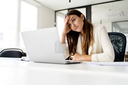 Photo for A businesswoman sits at the office desk, her head resting on her hand in a gesture of fatigue or frustration, a relatable moment for many professionals facing challenging tasks or long work hours - Royalty Free Image