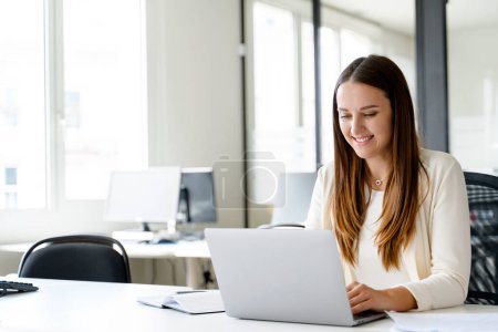 Une jeune femme d'affaires est capturée dans un moment de joie sincère alors qu'elle travaille sur son ordinateur portable, assise à son bureau. Aspects agréables de la vie au bureau et la satisfaction qui découle de l'engagement au travail.