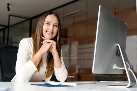 Foto de Joven mujer de negocios mirando a la cámara con una sonrisa amigable, usando la computadora en una oficina brillante, su sonrisa sugiriendo facilidad y competencia con la tecnología. Trabajadora de oficina en el lugar de trabajo - Imagen libre de derechos